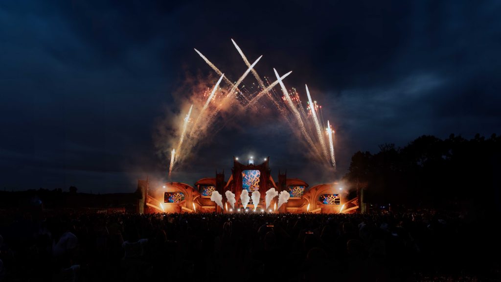 Auf dem Bild ist die Mainstage des Festivals OpenBeatz. Die Bühne ist orange beleuchtet, gerade kommen Flammen aus der Bühne und im Hintergrund ist Feuerwerk.
